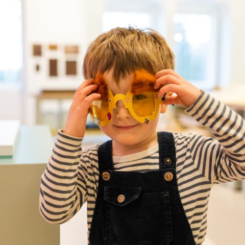 A young child wearing striped long sleeves and dark overalls smiles while holding up large, playful yellow sunglasses decorated with orange fuzzy eyebrows. The child stands indoors in a brightly lit room with soft-focus windows and displays in the background.