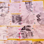 A grid of small, monochrome printed art pieces is arranged on a bright yellow background. Each piece shows various textures and patterns, including leaves, feathers, geometric shapes, and bubble-wrap impressions. The papers are connected with colourful plastic links in orange, pink, and blue.