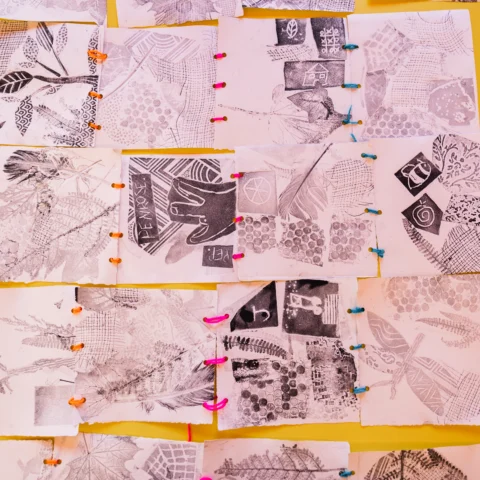 A grid of small, monochrome printed art pieces is arranged on a bright yellow background. Each piece shows various textures and patterns, including leaves, feathers, geometric shapes, and bubble-wrap impressions. The papers are connected with colourful plastic links in orange, pink, and blue.