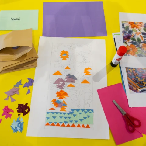 A brightly coloured craft table covered with paper scraps, envelopes labeled with colors, a glue stick, scissors, and printed floral reference images. In the center is a drawing in progress being decorated with small triangular pieces of coloured paper arranged in patterns.