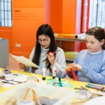 Two children sit at a brightly colored craft table in an orange-walled room, working on art projects. The child on the left, wearing glasses and a light gray sweater, holds scissors and a piece of paper. The child on the right, wearing a light blue sweatshirt, handles a piece of red translucent material. Various craft supplies—paper, glue sticks, sticks, tape, and markers—are scattered across the table.
