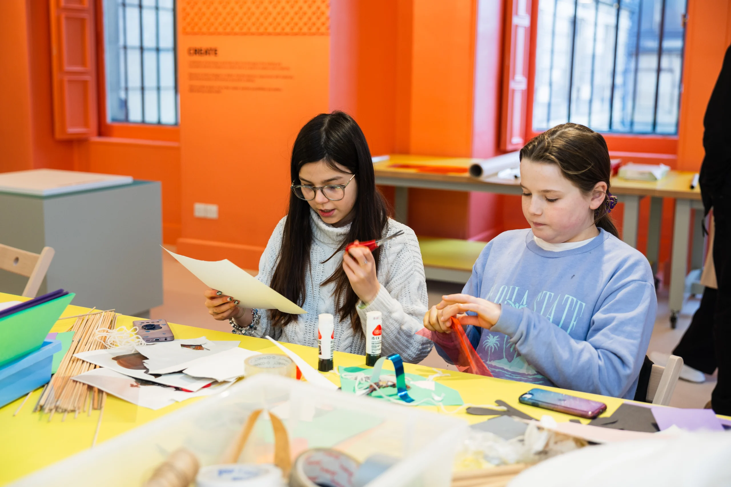 Two children sit at a brightly colored craft table in an orange-walled room, working on art projects. The child on the left, wearing glasses and a light gray sweater, holds scissors and a piece of paper. The child on the right, wearing a light blue sweatshirt, handles a piece of red translucent material. Various craft supplies—paper, glue sticks, sticks, tape, and markers—are scattered across the table.