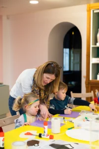 A woman leans over a table helping two young children with an arts-and-crafts activity. The children sit at a bright yellow table covered with paper, glue sticks, markers, string, and other craft supplies. The girl on the left is drawing on purple paper while the boy on the right works with a marker. The scene takes place in a well-lit room with pastel walls and wooden shelves in the background.