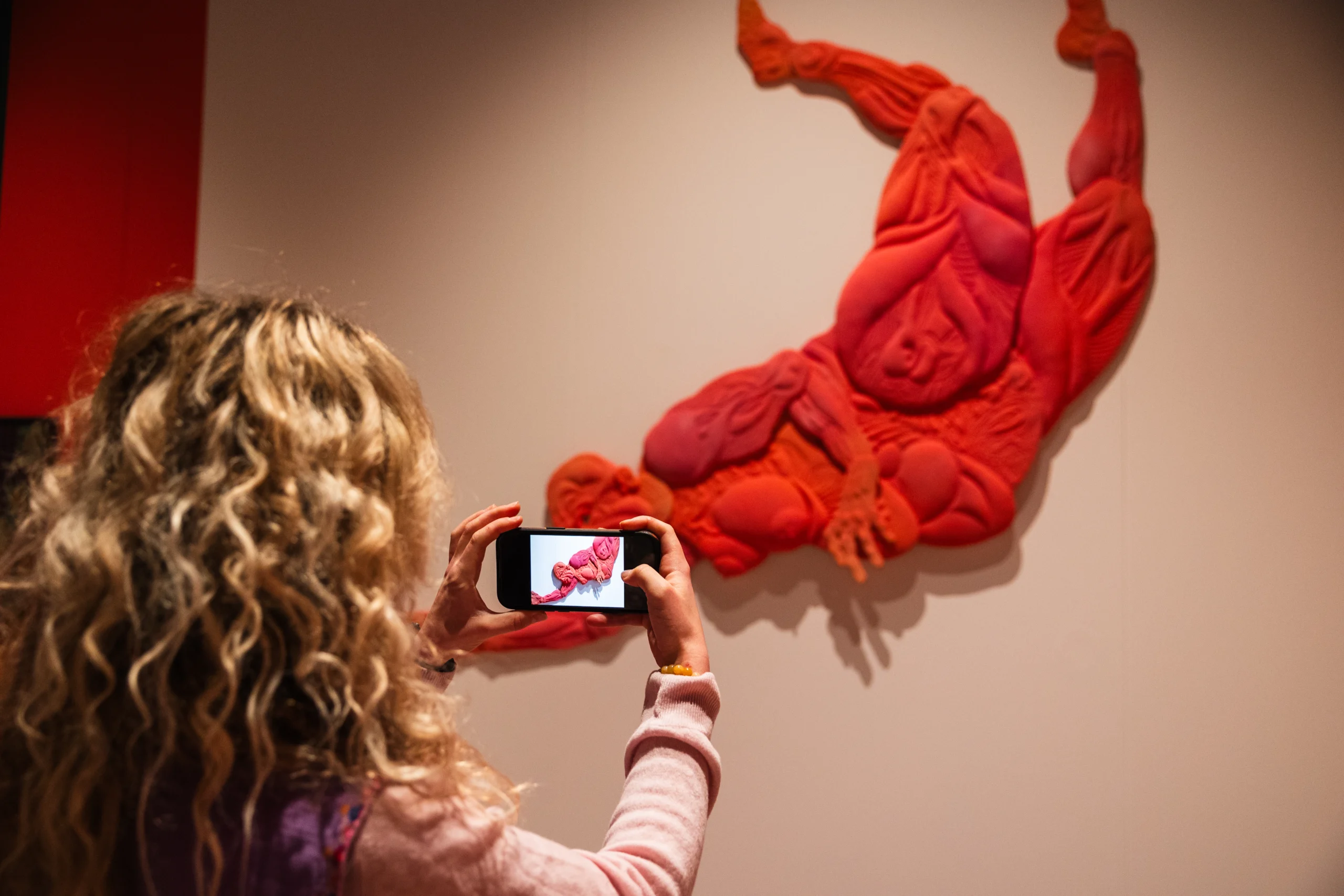 A person with long curly hair is photographing a vibrant red and orange wall-mounted sculpture using a smartphone. The artwork depicts an abstract, contorted human figure with exaggerated muscular forms and textured surfaces, creating a dynamic sense of movement. The background wall is neutral, emphasizing the bold colors and intricate details of the piece.