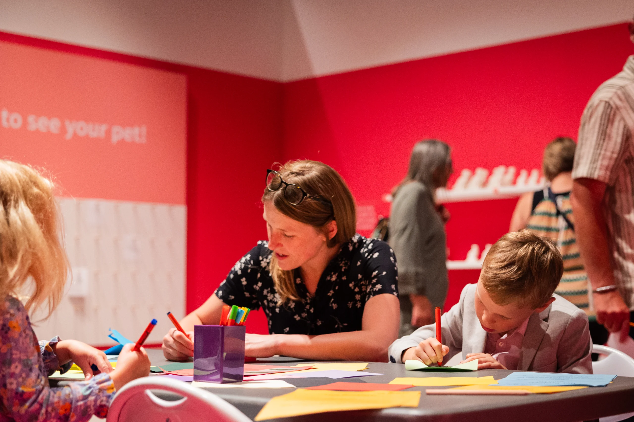 A group of people seated at a table in a brightly lit room with red walls, engaged in an arts and crafts activity. The table is covered with colorful paper sheets, markers, and other craft supplies. In the background, more individuals are visible near a display wall with small white objects arranged in rows.