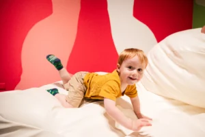 A young child with light hair, wearing a mustard-yellow shirt, khaki shorts, and colorful socks, smiles while crawling on a large white cushioned surface in a brightly colored play area with red and pink abstract shapes on the wall behind.