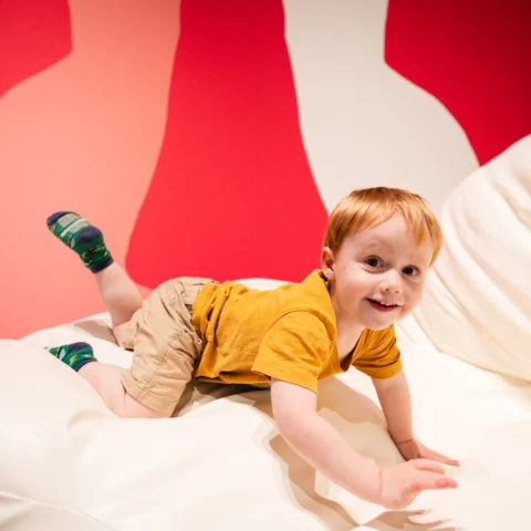 A young child with light hair, wearing a mustard-yellow shirt, khaki shorts, and colorful socks, smiles while crawling on a large white cushioned surface in a brightly colored play area with red and pink abstract shapes on the wall behind.