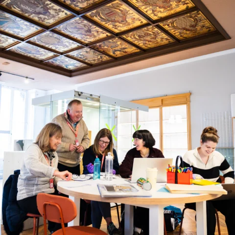 A group of five people seated and standing around a round table in a bright gallery space with wooden floors and large windows. The table is covered with laptops, notebooks, colorful stationery, and water bottles, suggesting a collaborative meeting or workshop. Above, an ornate ceiling panel features decorative artwork in rich tones, adding a historic element to the modern setting.