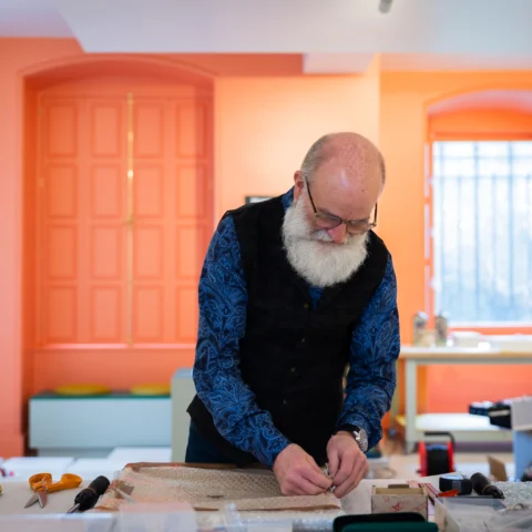 A person working at a table in a brightly lit room with orange walls and large windows. The individual is focused on a craft or restoration task, handling a piece of fabric or material on the tabletop. Surrounding the workspace are tools, small boxes, and equipment, suggesting a hands-on project in progress.
