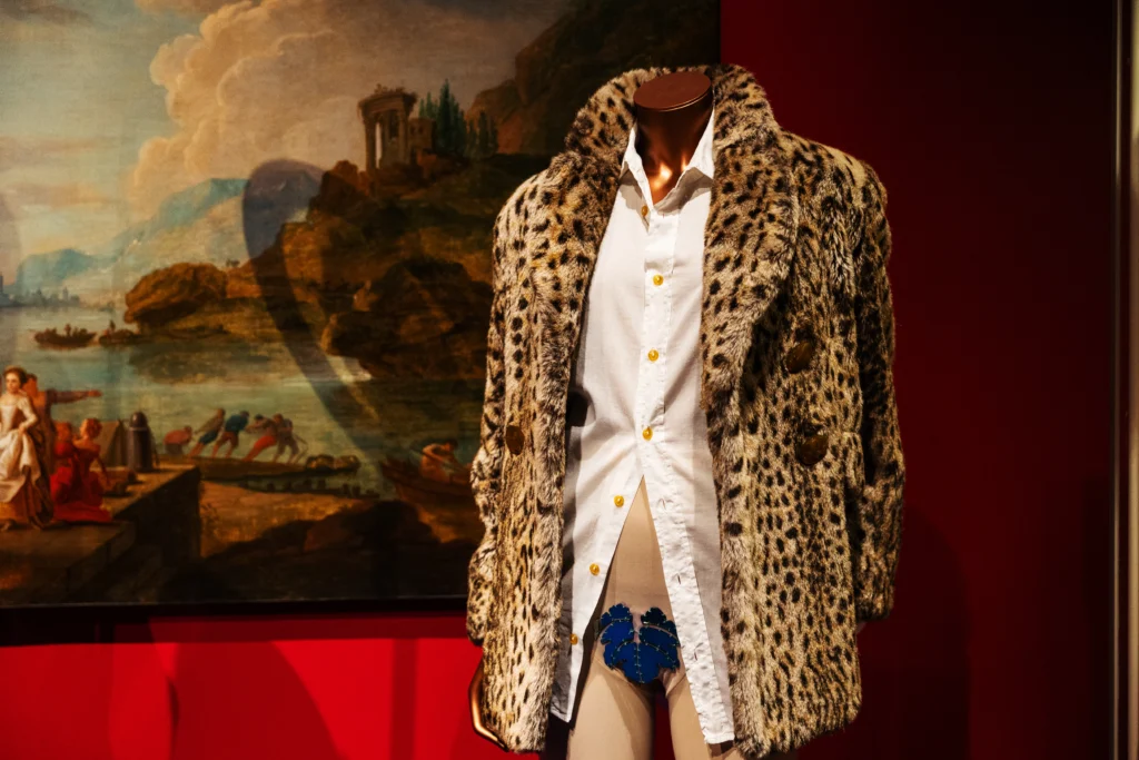 A mannequin is dressed in a leopard‑patterned faux‑fur coat worn over a white shirt with gold buttons. The shirt is slightly unbuttoned at the lower edge, revealing a decorative blue undergarment. Behind the mannequin is a large classical landscape painting.