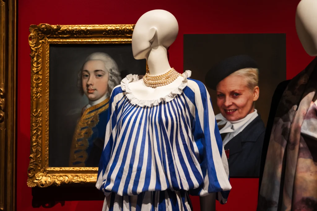 A mannequin wearing a blue‑and‑white vertically striped blouse with a ruffled neckline is displayed against a deep red gallery wall. The mannequin’s outfit is accessorised with layered pearl necklaces. Behind the mannequin hang framed portraits in ornate gold frames.