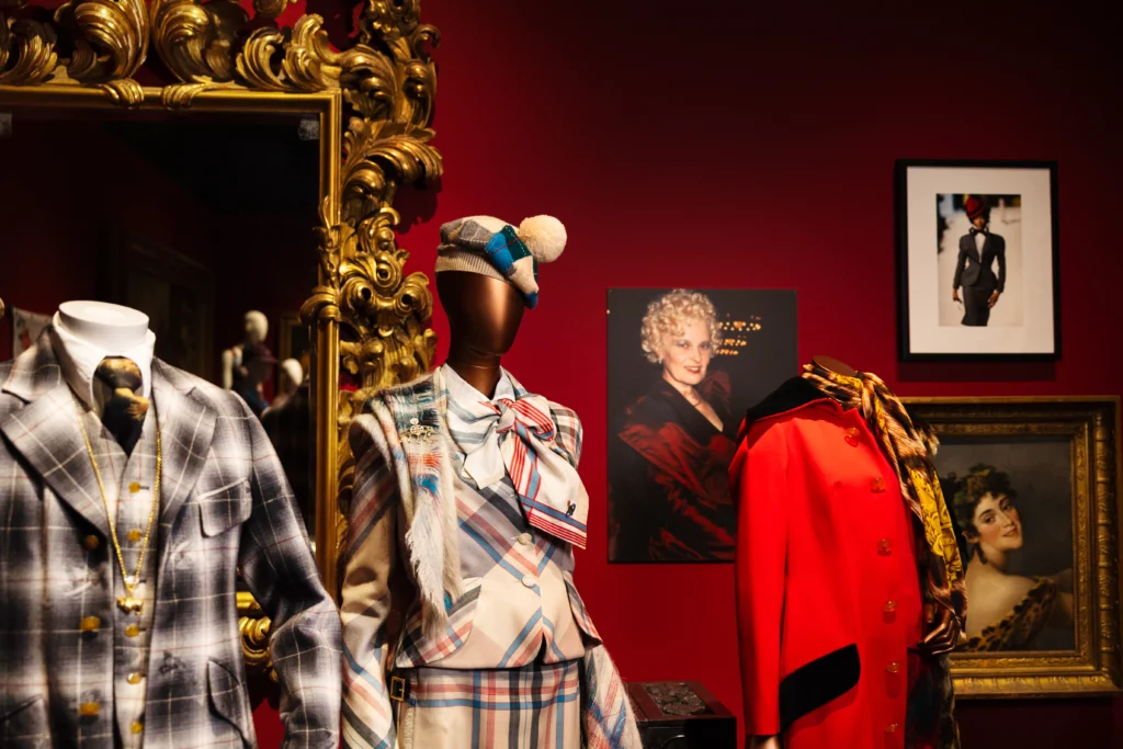 Mannequins dressed in tailored garments stand among ornate framed artworks and an elaborate gilded mirror. The outfits include plaid suits and a coordinating checked ensemble with a bow and hat. A bright red coat is displayed on another mannequin, accompanied by additional framed imagery on the wall.