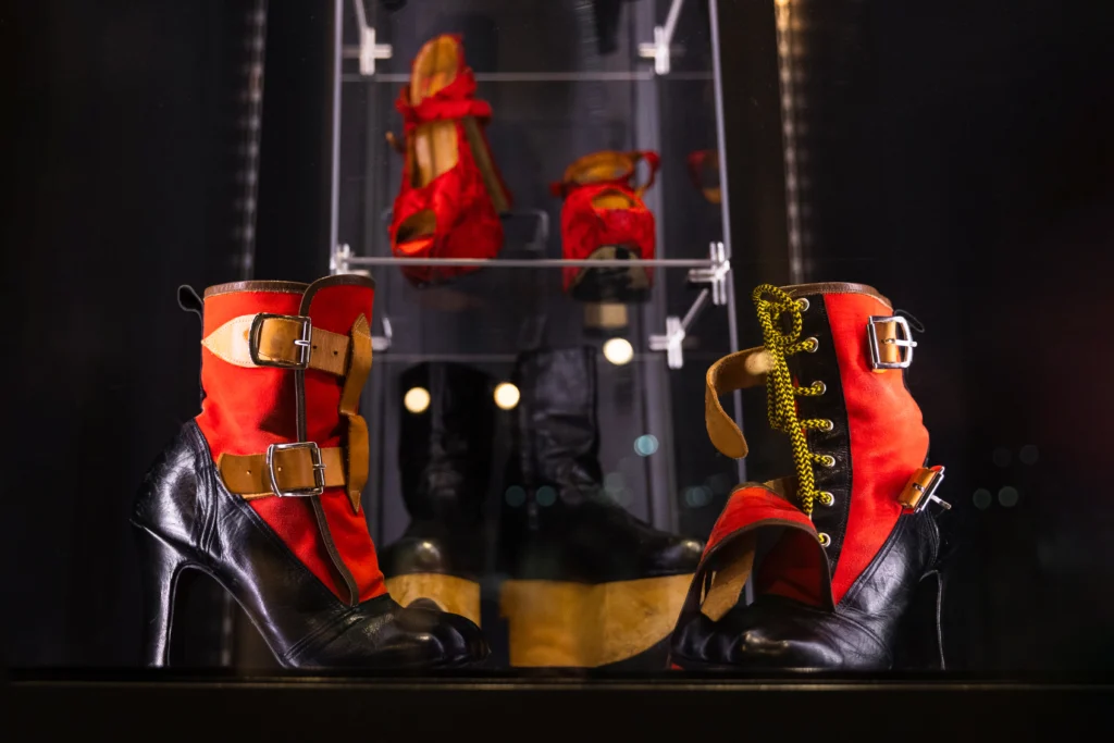 A pair of heeled boots is displayed in a glass case. The boots combine black leather with red fabric panels and feature multiple metallic buckles and straps. One boot also has gold-toned lacing running up the front. Reflections of additional footwear, including red platform shoes, are visible in the background.