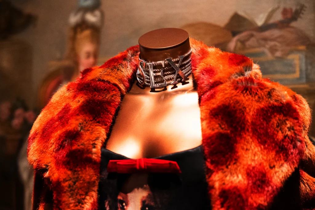 A mannequin wears a vibrant red‑and‑orange fur coat draped over a dark garment with a small red bow at the neckline. Around the mannequin’s neck is a prominent metallic choker featuring large block‑letter forms. A historic-style painting forms the backdrop.