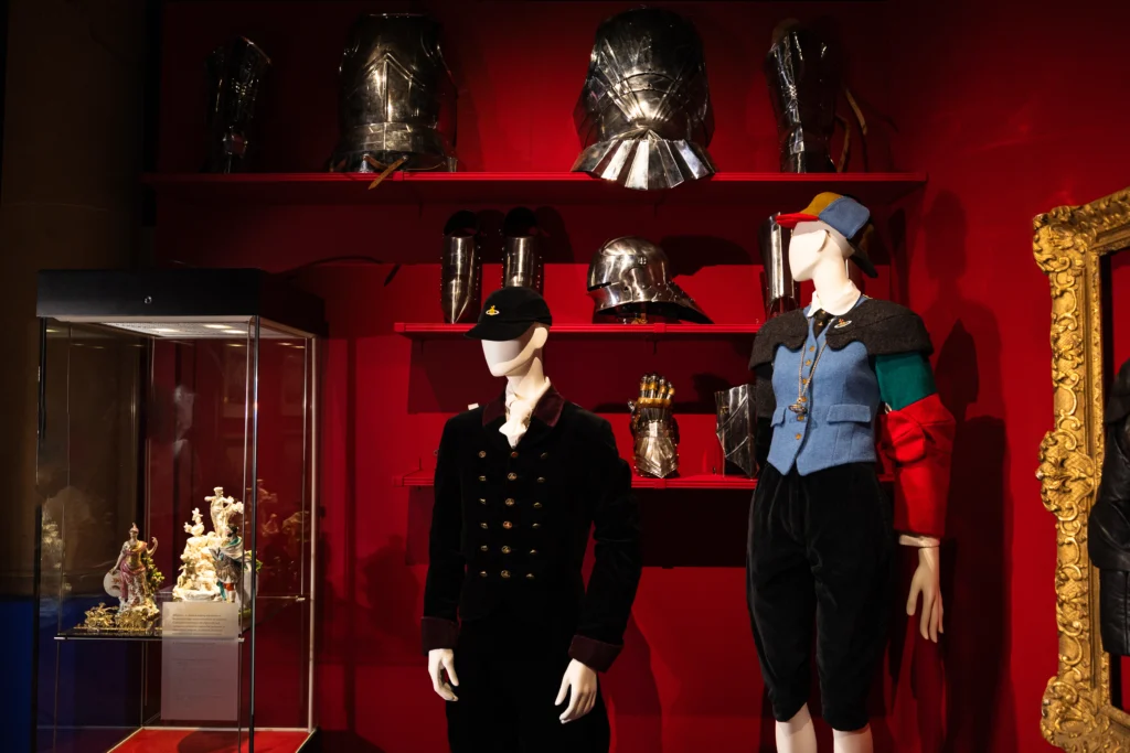Two mannequins are displayed in front of a deep red wall. One mannequin wears a dark tailored outfit with metallic buttons and a cap. The other wears a colourful ensemble with red, blue, and green elements and a matching cap. Shelves above them hold polished pieces of armour, including helmets and a gauntlet. A glass case to the side contains decorative ceramic figures.