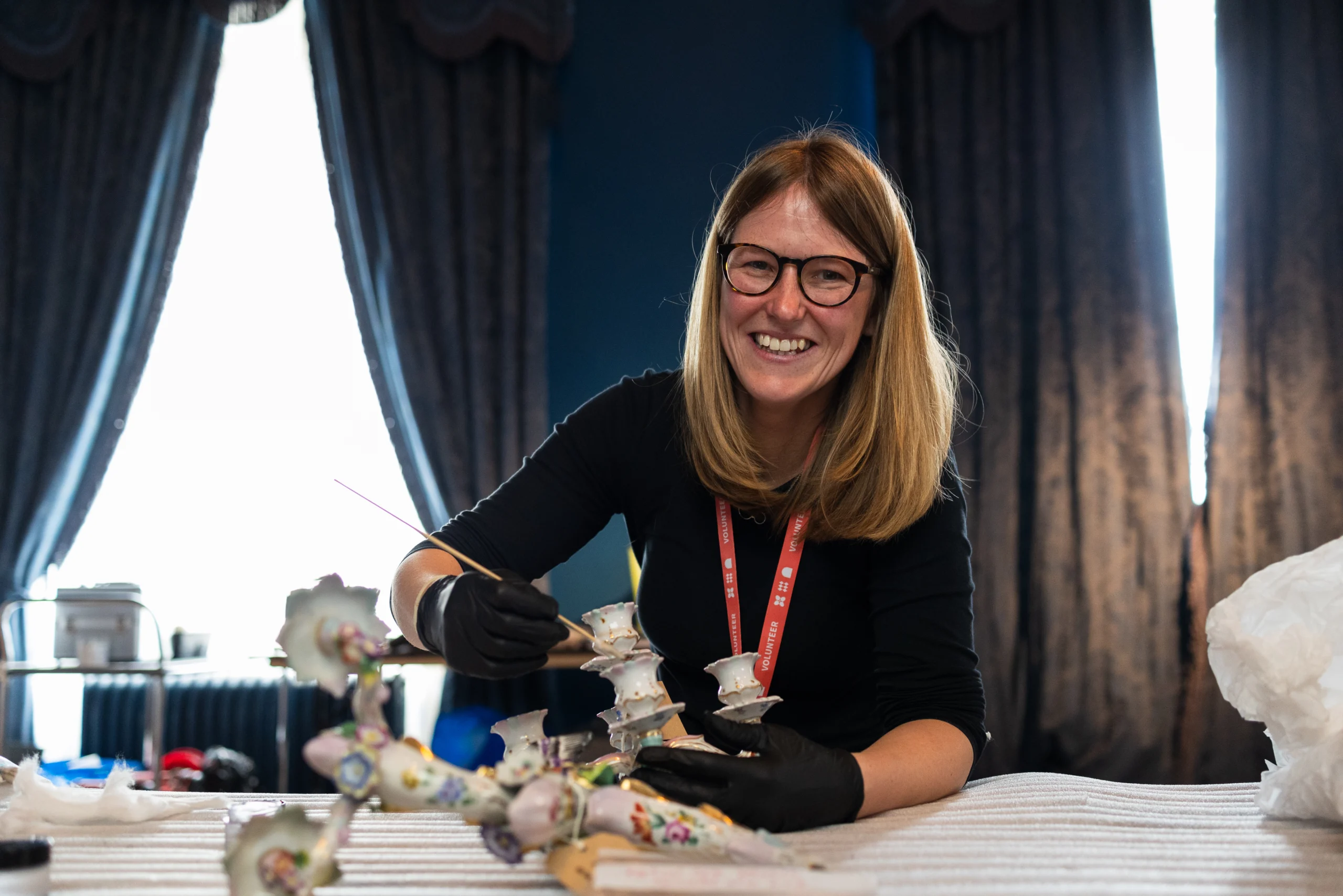 A person wearing black gloves is carefully restoring or cleaning a decorative porcelain piece on a table covered with a white cloth. The porcelain object features intricate floral patterns and pastel colors, with multiple small components arranged in front. The individual is using a fine brush and appears to be working in a room with dark curtains and large windows that allow natural light to illuminate the workspace.