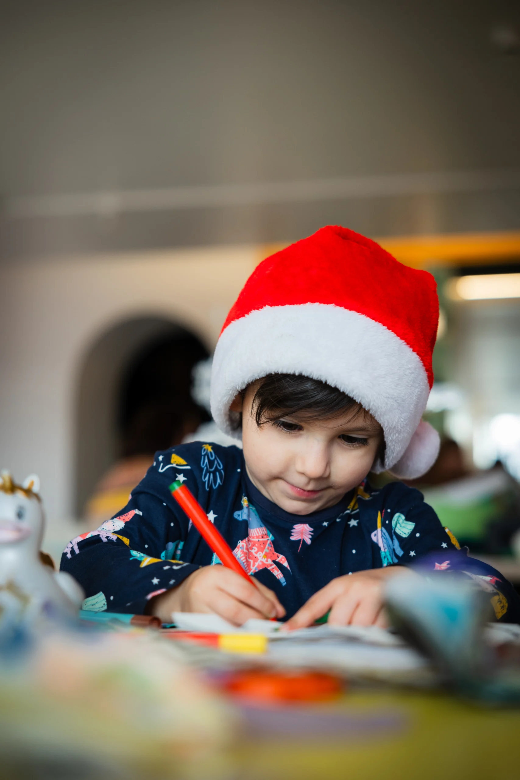 A child wearing a red Santa hat and a dark long-sleeved shirt with colorful patterns is drawing with a red pencil at a table. There are scattered papers and art supplies on the table, and a small toy unicorn is visible in the foreground. The background shows an indoor setting with blurred details.