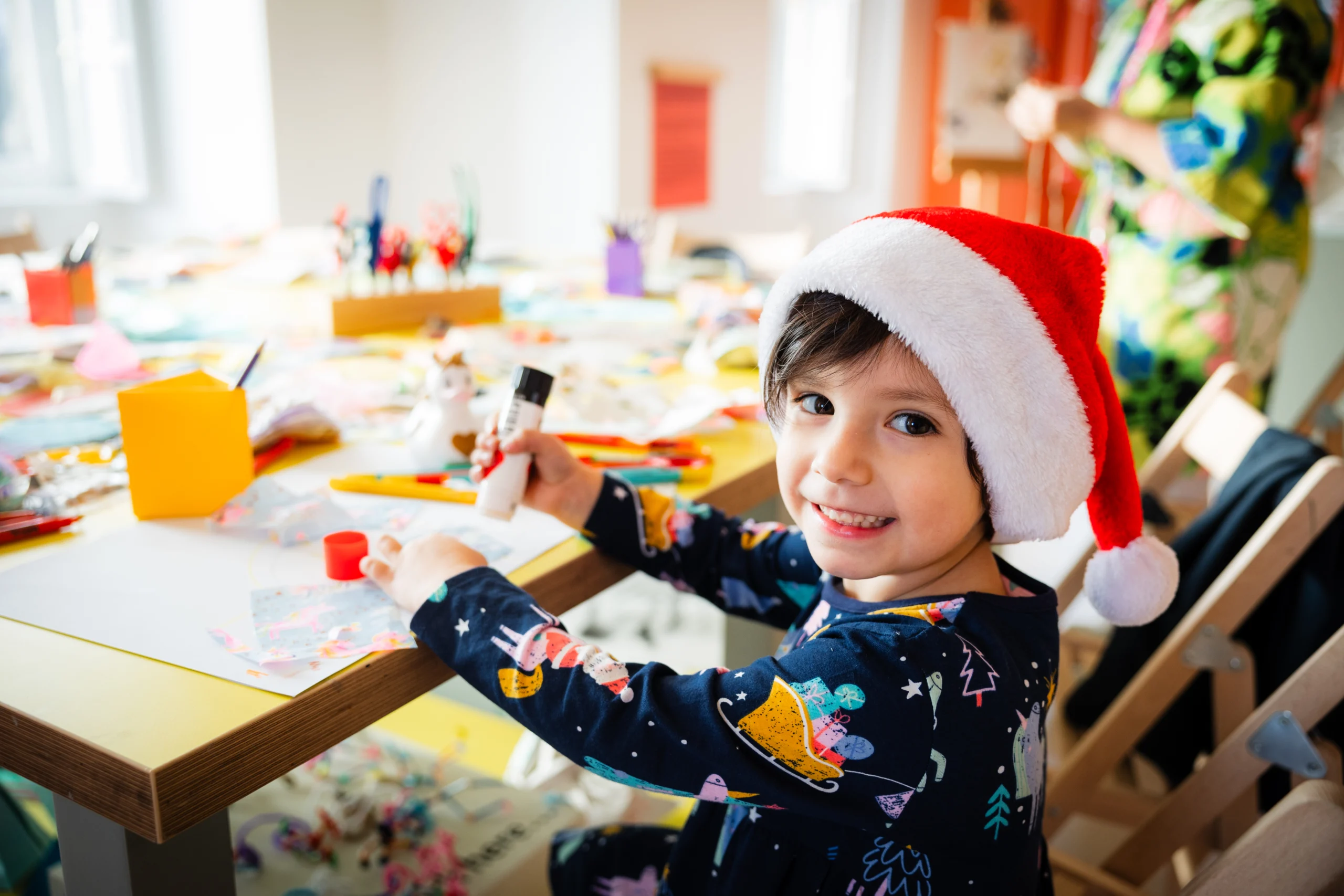 A young child wearing a Santa hat smiles at the camera while doing arts and crafts at a colorful, busy table covered with paper, markers, and craft supplies. The child holds a glue stick and appears to be working on a festive project in a bright, cheerful room.