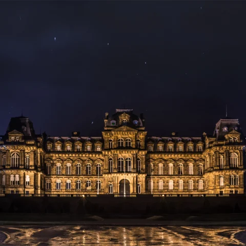 A grand historic building is illuminated at night with warm golden lighting, highlighting its ornate architecture and symmetrical façade. The dark sky above is scattered with faint star trails, and the building’s reflection glimmers in the water in the foreground.
