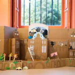 A large carboard version of The Bowes Museum with arched windows, paper flowers, small cut-out figures, and miniature cardboard cars, displayed indoors in front of an open orange-framed window.