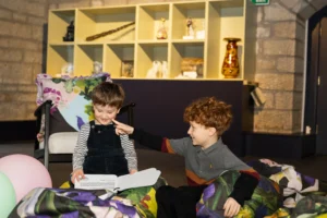 Two young children sit together indoors on colorful cushions. One child with brown hair, wearing striped sleeves and dark overalls, smiles while holding an open book. The other child with curly red hair gently touches the first child’s cheek, smiling playfully. Behind them is a shelving display with various decorative objects and vases.