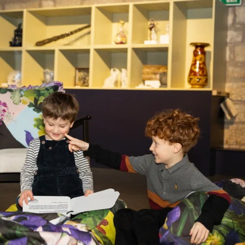 Two young children sit together indoors on colorful cushions. One child with brown hair, wearing striped sleeves and dark overalls, smiles while holding an open book. The other child with curly red hair gently touches the first child’s cheek, smiling playfully. Behind them is a shelving display with various decorative objects and vases.