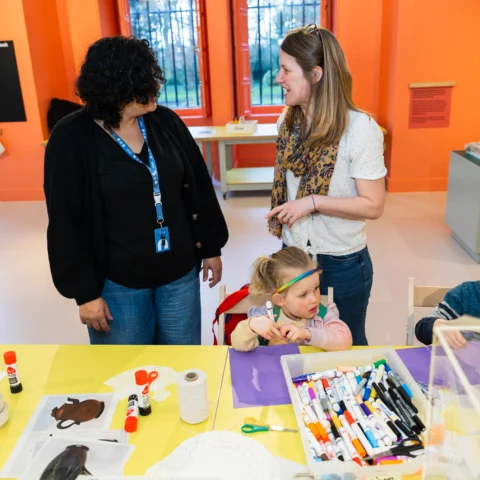 In the Create Space, a brightly coloured room where two adults stand talking while two young children sit at a table doing arts and crafts. The table is covered with markers, glue sticks, scissors, paper, and printed images. One child concentrates on a piece of paper, while the other holds a marker. The room has orange walls, large windows, and museum-style displays in the background.