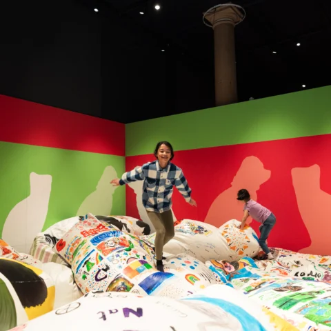 Two children play on a large, soft, mound-like installation covered in colourful drawings and patterns. One child balances with arms outstretched while another climbs nearby. The room has bold pink and green walls with silhouetted figures painted on them, and a tall column rises to a dark ceiling with spotlights, suggesting a museum or gallery setting.