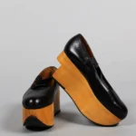 A pair of black leather platform shoes with very thick, light-colored wooden soles. One shoe is upright while the other leans against it, showing the exaggerated height and chunky shape of the platforms.