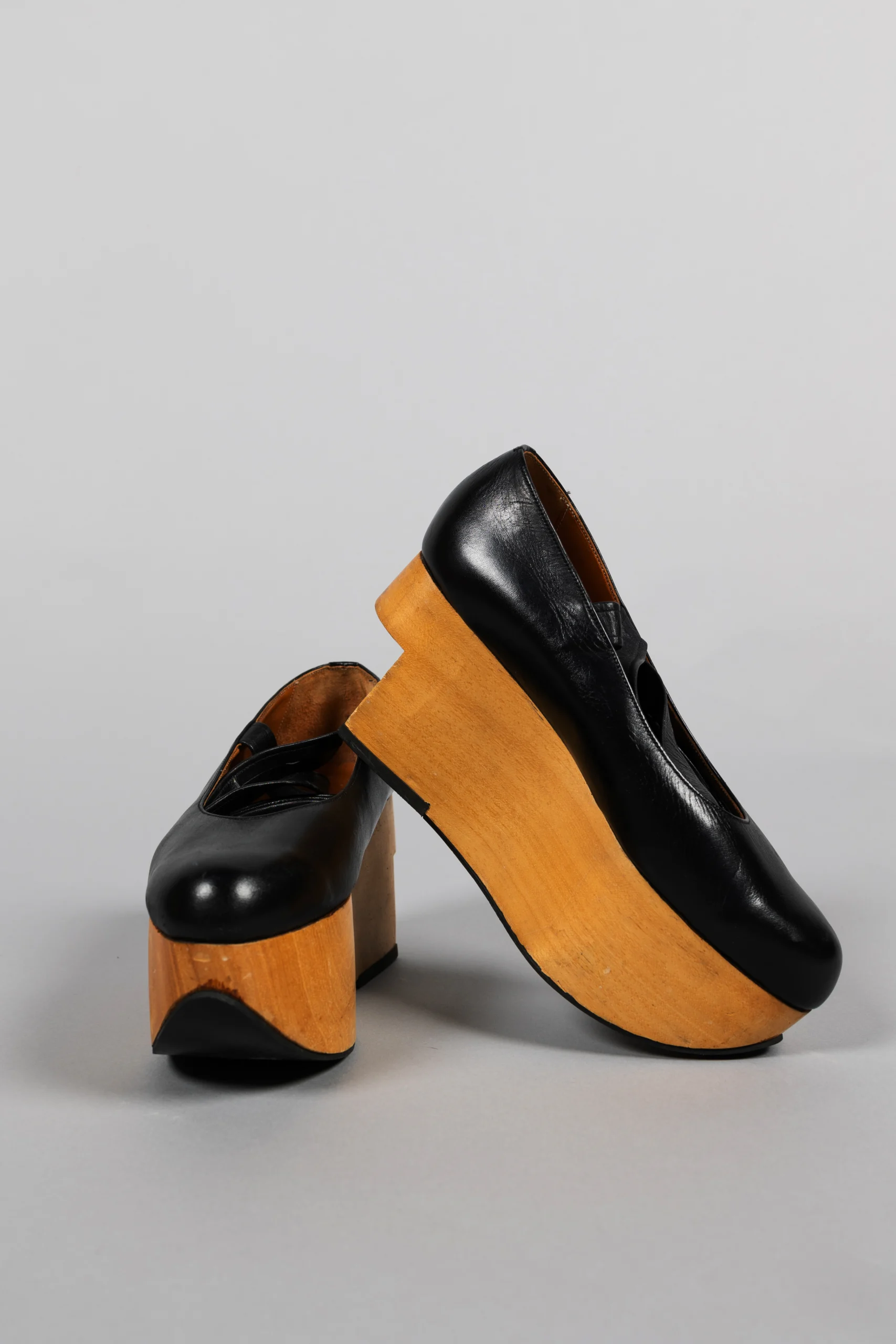 A pair of black leather platform shoes with very thick, light-colored wooden soles. One shoe is upright while the other leans against it, showing the exaggerated height and chunky shape of the platforms.