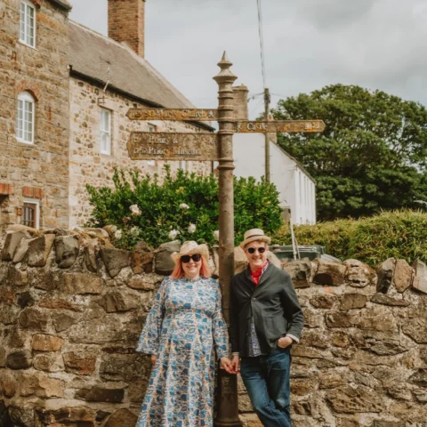 Founders of For the Love of the North Paul and Lucy Hull stand hand-in-hand beside an old stone wall and a vintage signpost in a quaint village. They wear hats and sunglasses, posing cheerfully in front of rustic stone buildings.