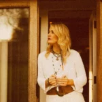 A person stands in a doorway holding a mug with both hands. They are wearing a light-coloured knit sweater, layered necklaces, bracelets, and a brown belt bag. Warm, golden lighting illuminates the wooden doorframe and the scene, giving it a cozy, relaxed atmosphere.