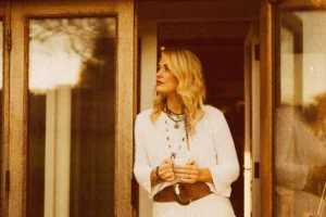 A person stands in a doorway holding a mug with both hands. They are wearing a light-coloured knit sweater, layered necklaces, bracelets, and a brown belt bag. Warm, golden lighting illuminates the wooden doorframe and the scene, giving it a cozy, relaxed atmosphere.