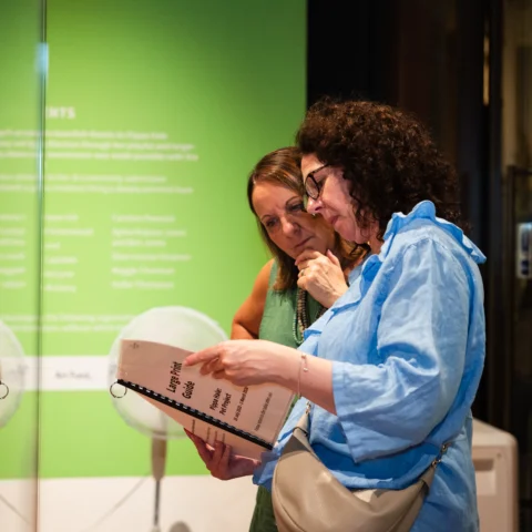 Two people stand together in a gallery space, looking at a large printed booklet or gallery guide. One person wearing a light blue shirt holds the guide open while pointing to the text. Behind them is a bright green exhibition wall with white text and reflections on the glass surface.