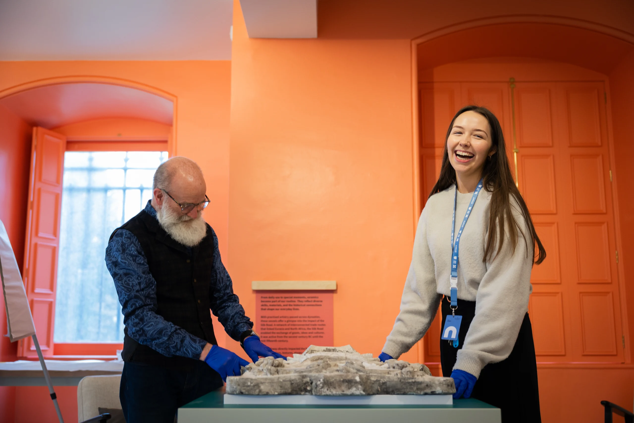 Two individuals standing at a table in a brightly lit room with orange walls and matching door panels. Both are wearing blue protective gloves and examining a large, irregularly shaped object that appears to be a stone or plaster artifact with textured surfaces. One person is wearing a dark patterned vest over a shirt, while the other has a light-colored sweater and a lanyard with an ID badge. A window with orange shutters and a flip chart are visible in the background, along with a wall-mounted information plaque.