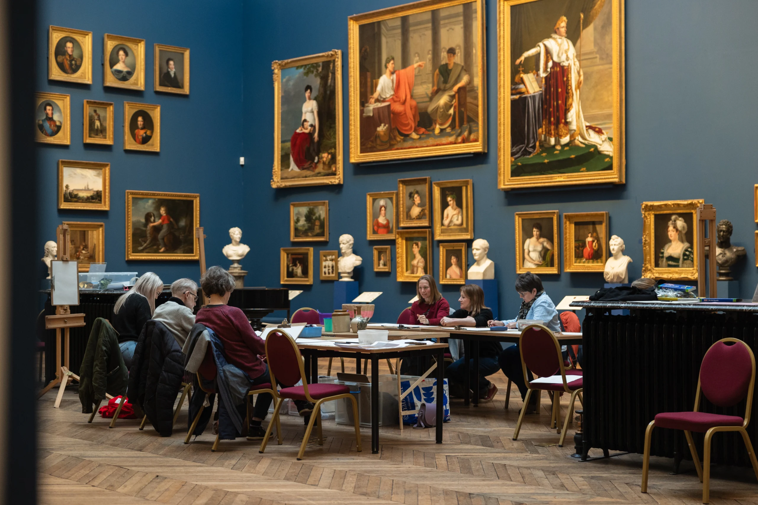 A group of people sit around tables in a large gallery room, participating in an art or study session. The space is surrounded by an extensive display of framed portraits and paintings in ornate gold frames, along with classical white busts arranged on shelves. The room has blue walls, wooden parquet flooring, and red chairs positioned around the tables. The atmosphere is calm and focused.