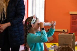 A child holds a homemade cardboard tube up to their eye like a telescope while standing at a craft table in a brightly coloured room with orange walls. The child wears a patterned headband and a smartwatch. On the table are craft supplies, including markers, scissors, and a cardboard box decorated with drawings. An adult stands nearby, partially visible.