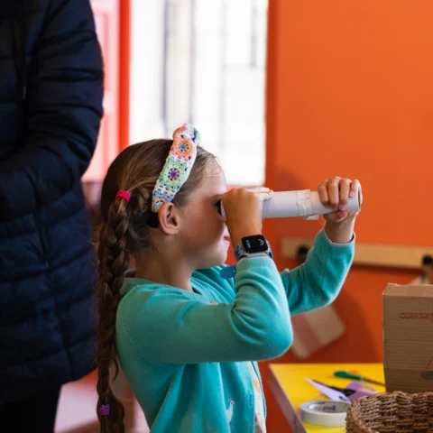 A child holds a homemade cardboard tube up to their eye like a telescope while standing at a craft table in a brightly coloured room with orange walls. The child wears a patterned headband and a smartwatch. On the table are craft supplies, including markers, scissors, and a cardboard box decorated with drawings. An adult stands nearby, partially visible.