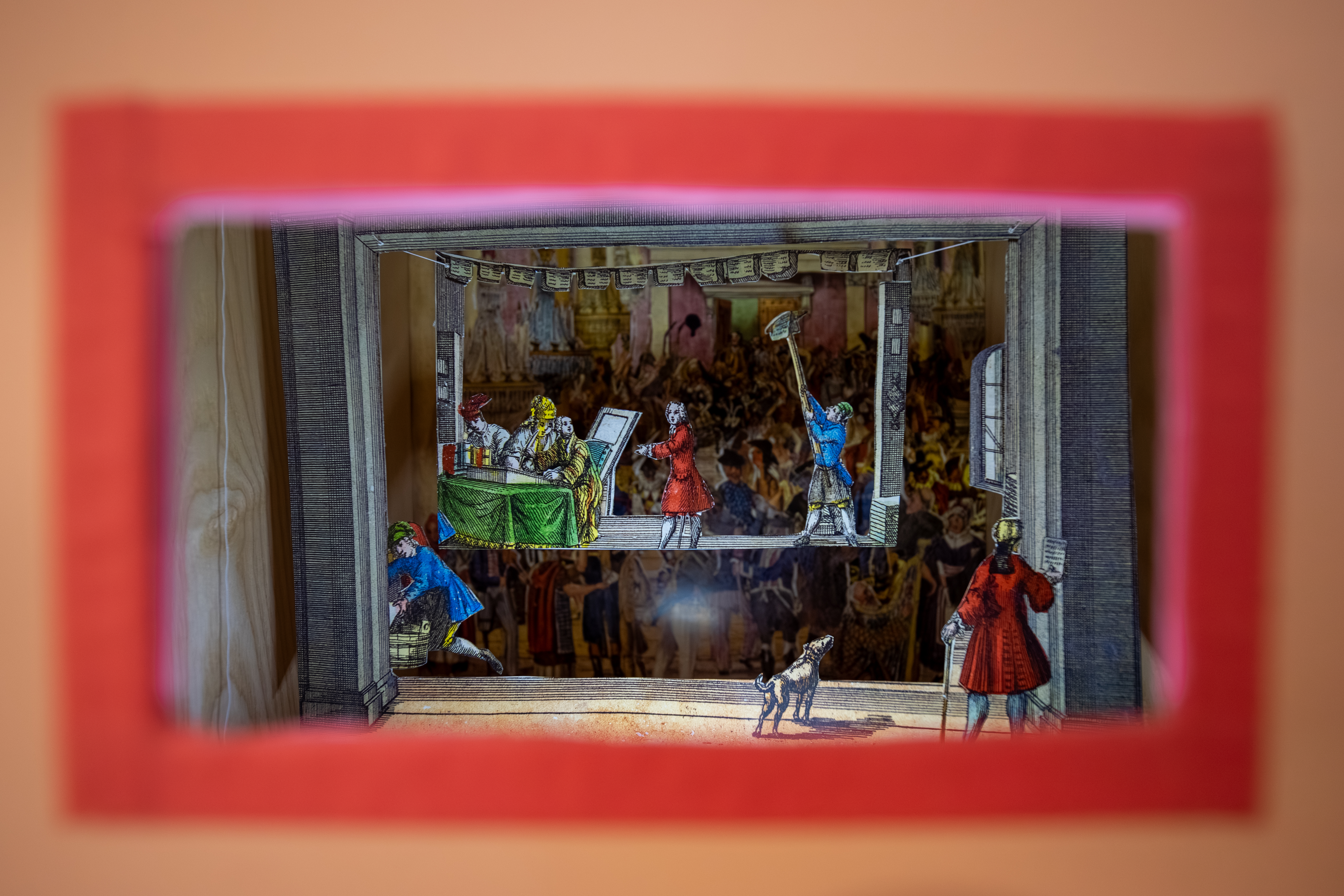 A brightly lit miniature theatre scene viewed through a red-framed window. The paper diorama shows several costumed figures on a stage, including performers holding props and a small dog at the front. A detailed illustrated audience forms the backdrop behind the stage.