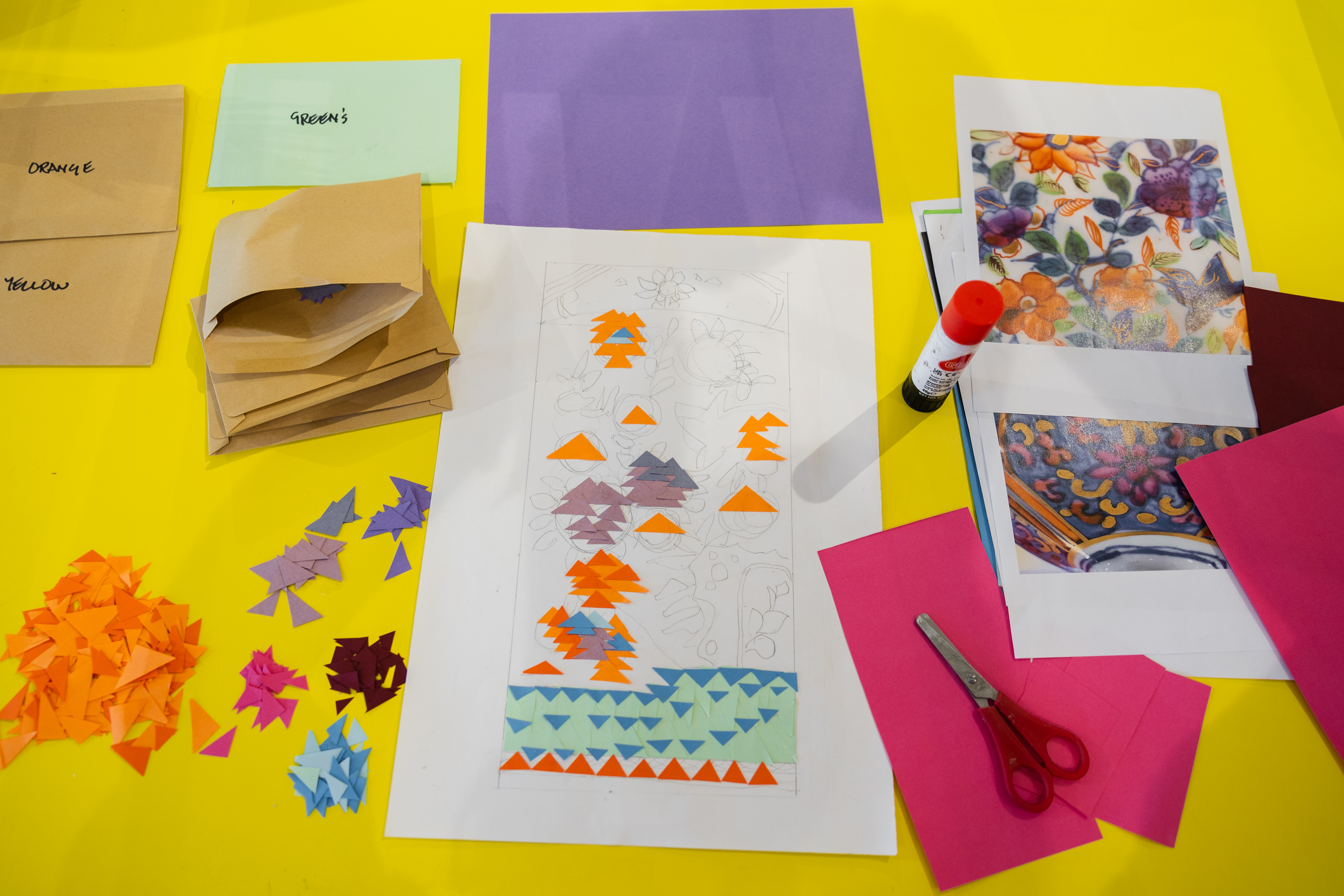 A bright yellow table covered with craft materials, including coloured paper, small paper shapes sorted by colour, envelopes labelled with colour names, a glue stick, scissors, and patterned reference sheets. At the centre is a drawing in progress decorated with colourful paper triangles.