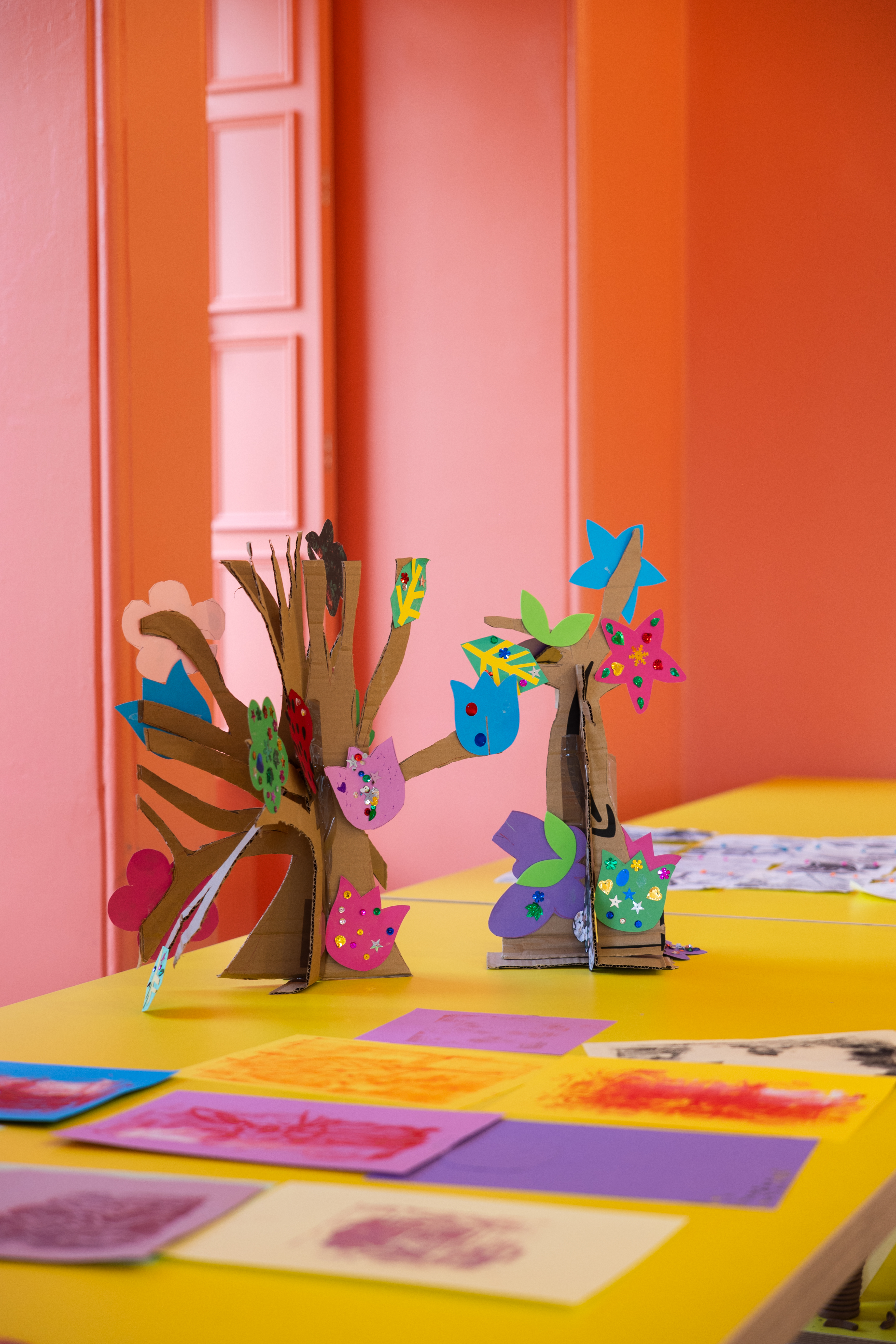 Colourful cardboard sculptures decorated with cut‑out paper shapes sit on a bright yellow table in an art room with orange and pink walls. Additional artwork made from painted and printed paper is laid out flat on the tabletop.