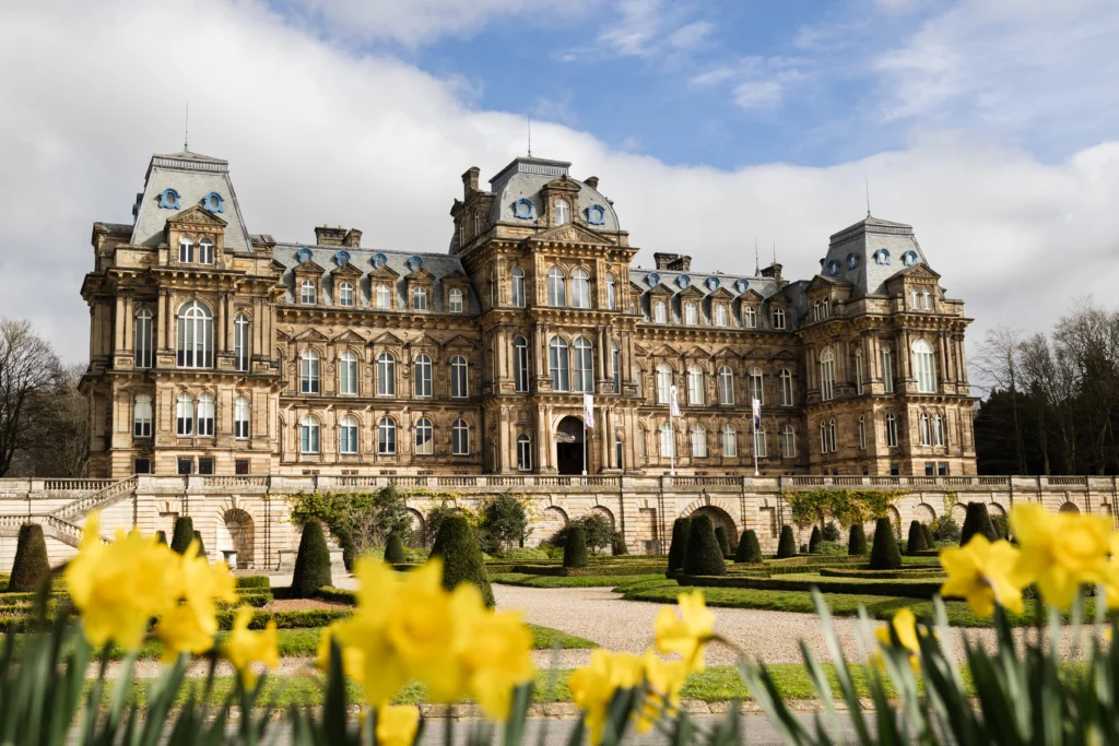 The Bowes Museum's stone architecture in front a manicured garden. The building features large windows, decorative roof gables and intricate detailing. In the foreground, bright yellow daffodils are in soft focus, framing the view of the building. The sky is blue and partly cloudy, creating a calm elegant atmosphere.