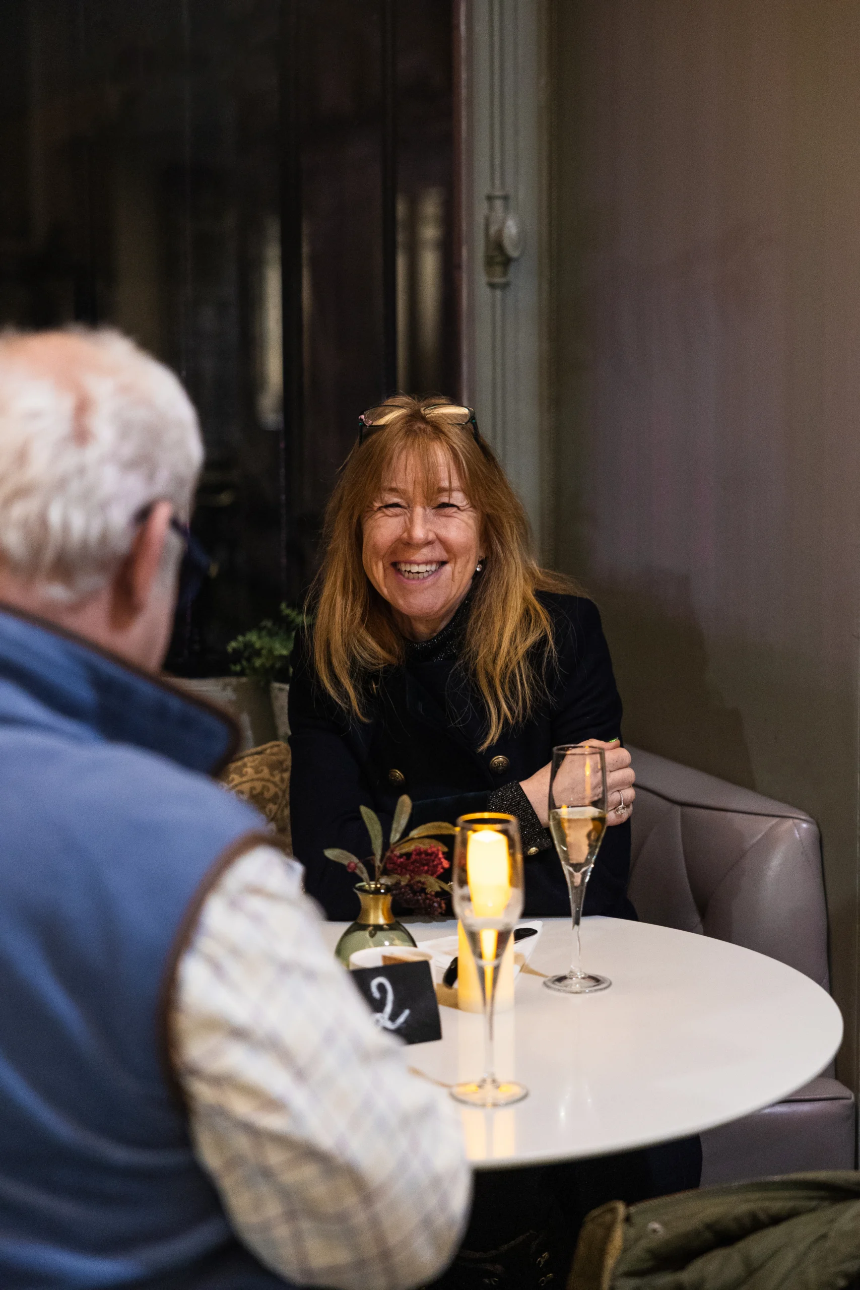 Two people sit at a small round table in a dimly lit café or bar. One person holds a glass of sparkling drink, and two illuminated candles sit on the table alongside a small vase with flowers and a table number sign.