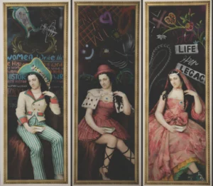 A triptych artwork featuring three life‑sized sculptural figures dressed in theatrical, stylised costumes, each seated within a tall framed display. The left figure wears striped trousers, a decorated jacket, and a large blue hat against a background covered in chalk‑like drawings and text. The central figure wears a pink patterned dress with a white, studded capelet, set before a dark backdrop filled with colourful doodles and symbols. The right figure wears a flowing pink gown with layered textures, seated in front of a background showing handwritten phrases including “LIFE HER LEGACY.” Each figure holds a small book or diary in one hand.