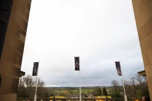 A view from beneath The Bowes Museum’s stone entrance archway looks out across the terrace and surrounding countryside. Three tall flagpoles stand in the foreground, each displaying a vertical exhibition banner featuring artworks and figures. Beyond the terrace balustrade are trees, rooftops, fields, and rolling hills under a wide grey sky. The stone architecture of the museum frames the scene on both sides.