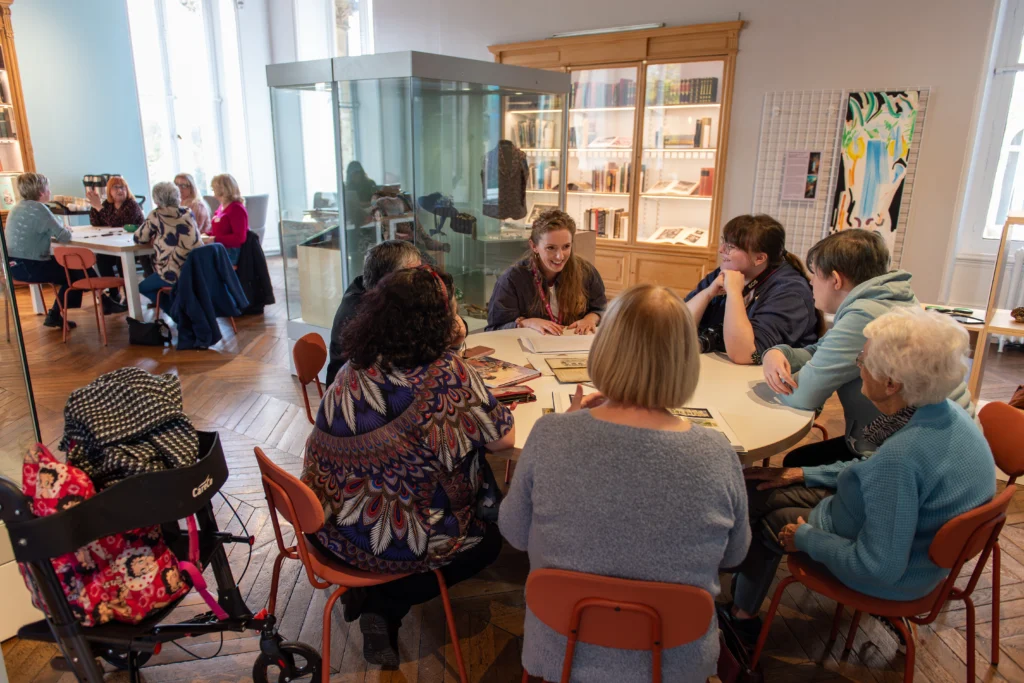 A group of people sit around tables in a bright museum space, participating in an activity involving art materials. Display cases with objects and books line the walls. The room has wooden, large windows and colourful artwork on the walls. A mobility aid with a floral bag is visible in the foreground beside one of the chairs.