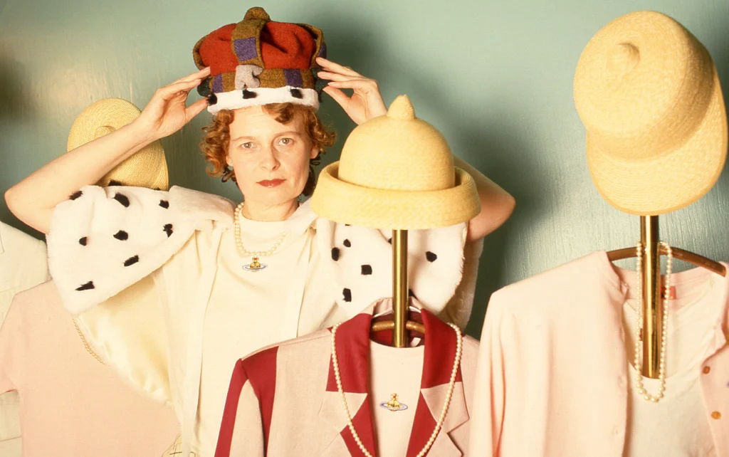 A person adjusts a colourful, oversized crown on their head while standing among mannequins dressed in pastel outfits with straw hats. They wear a cream outfit with a spotted, cape‑like accessory, creating a playful, theatrical scene.