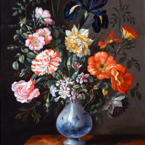 A painted still life shows a large bouquet of flowers arranged in a blue‑and‑white ceramic vase, set on a wooden tabletop. The bouquet includes roses, poppies, irises, carnations, and small daisy‑like flowers in shades of pink, red, orange, yellow, white, and deep blue, with green leaves and stems spreading outward. The background is dark and plain, creating contrast with the brightly coloured flowers and the reflective surface of the vase.