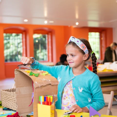 A child sits at a bright yellow craft table in a colourful room with orange walls and large windows. The child is creating a cardboard craft structure, using materials such as coloured pens, string, paper shapes, and glue. The table is covered with additional craft supplies, including scissors, tape, and paper tubes. An adult stands nearby, partially visible.