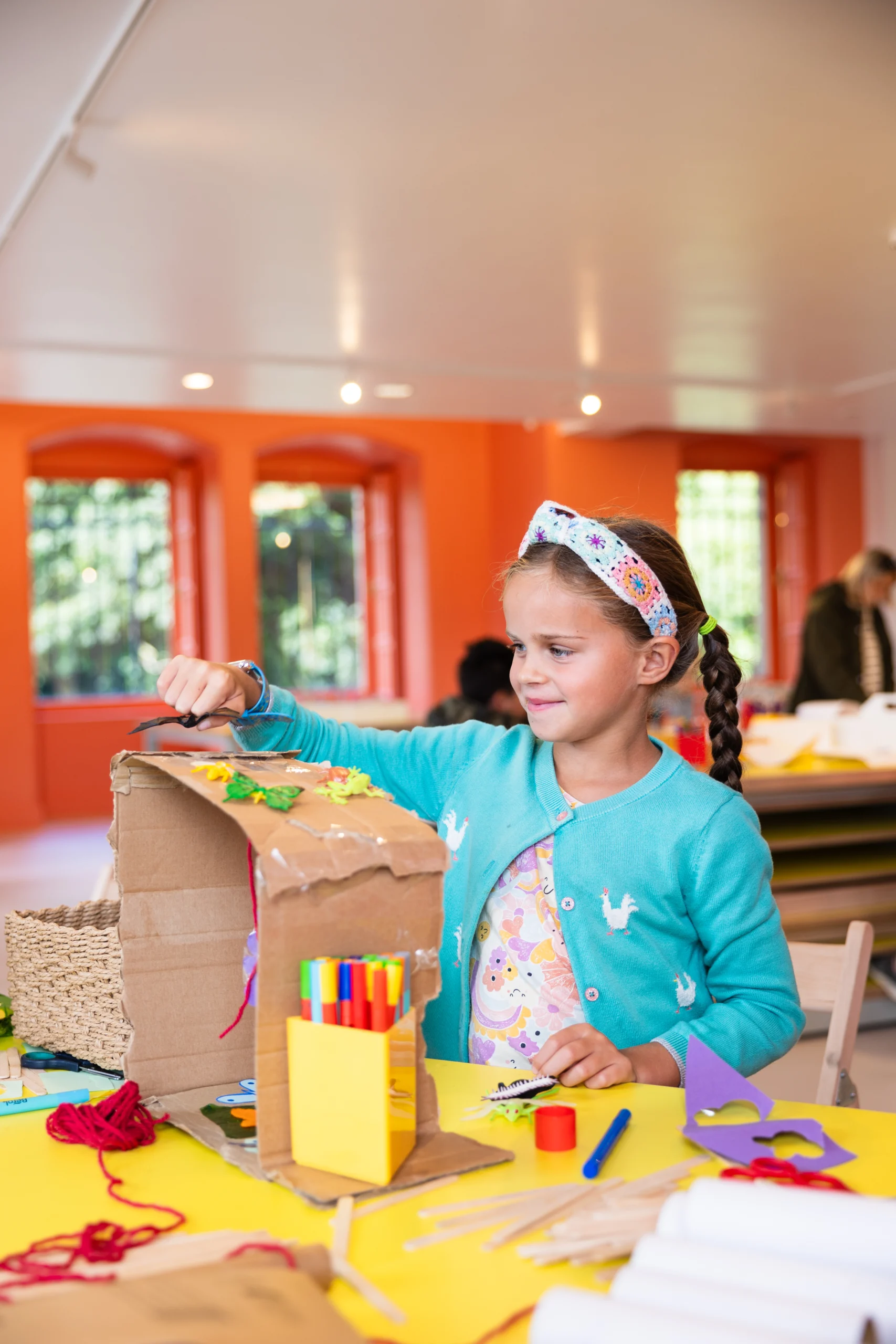 A child sits at a bright yellow craft table in a colourful room with orange walls and large windows. The child is creating a cardboard craft structure, using materials such as coloured pens, string, paper shapes, and glue. The table is covered with additional craft supplies, including scissors, tape, and paper tubes. An adult stands nearby, partially visible.
